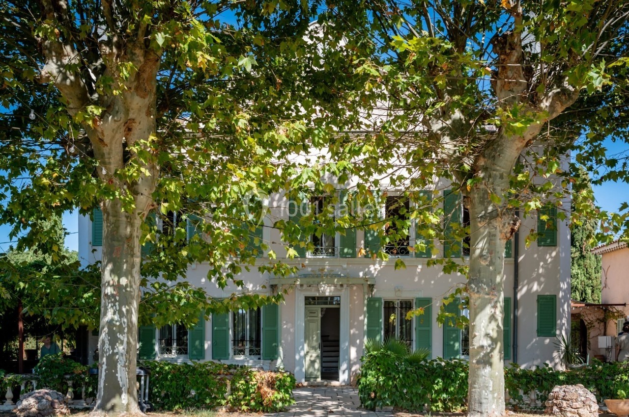 Façade d'une maison traditionnelle entourée de deux grands arbres avec des volets verts et un chemin d'entrée pavé.