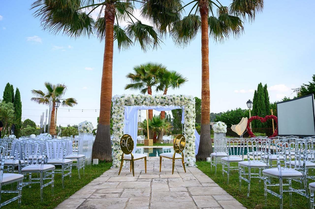 Décor de cérémonie en plein air avec arche fleurie, chaises transparentes et palmiers en arrière-plan.
