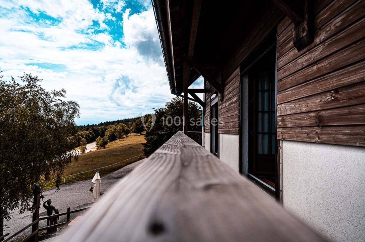 Vue depuis un balcon en bois d'une maison, donnant sur un paysage naturel avec arbres, prairie et ciel partiellement nuageux.
