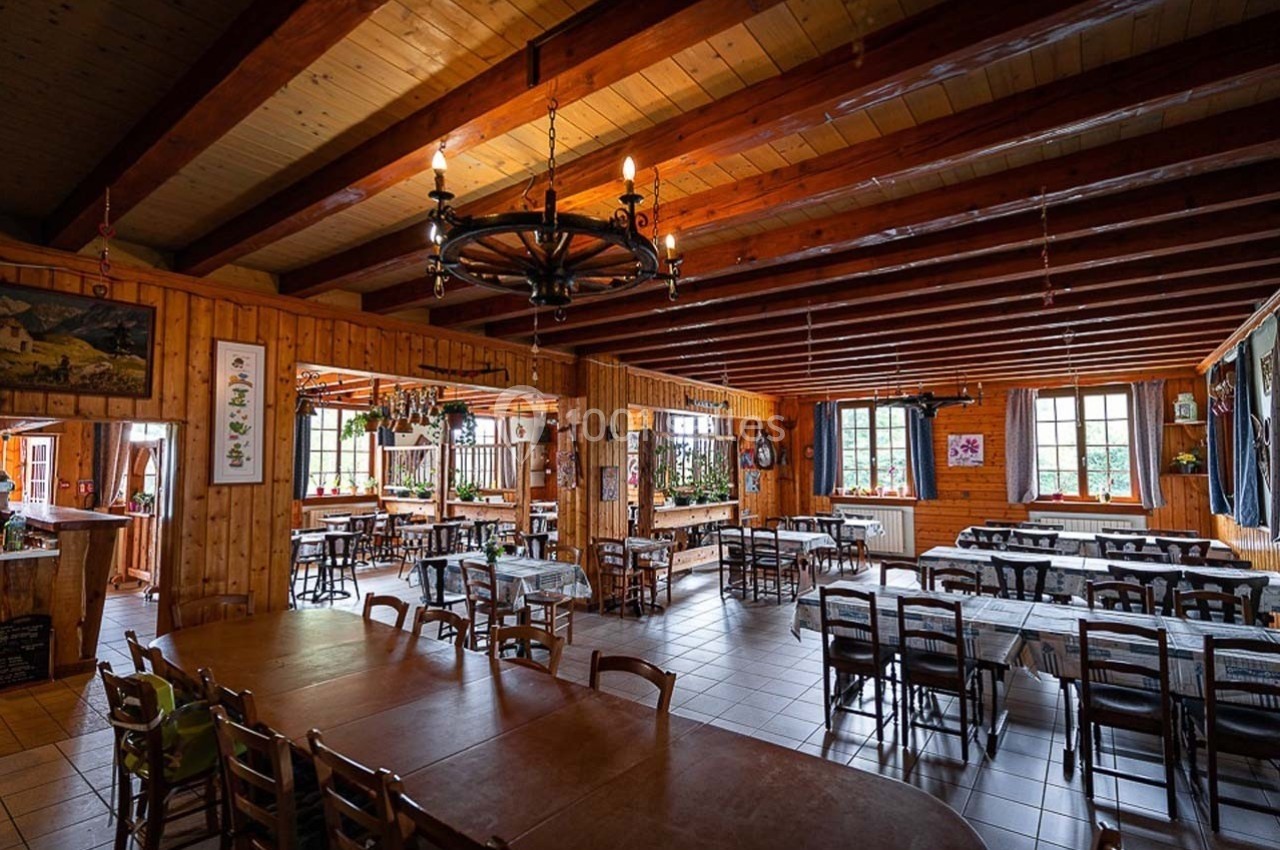 Salle à manger spacieuse en bois avec tables et chaises alignées, éclairée par des fenêtres et des lustres rustiques.