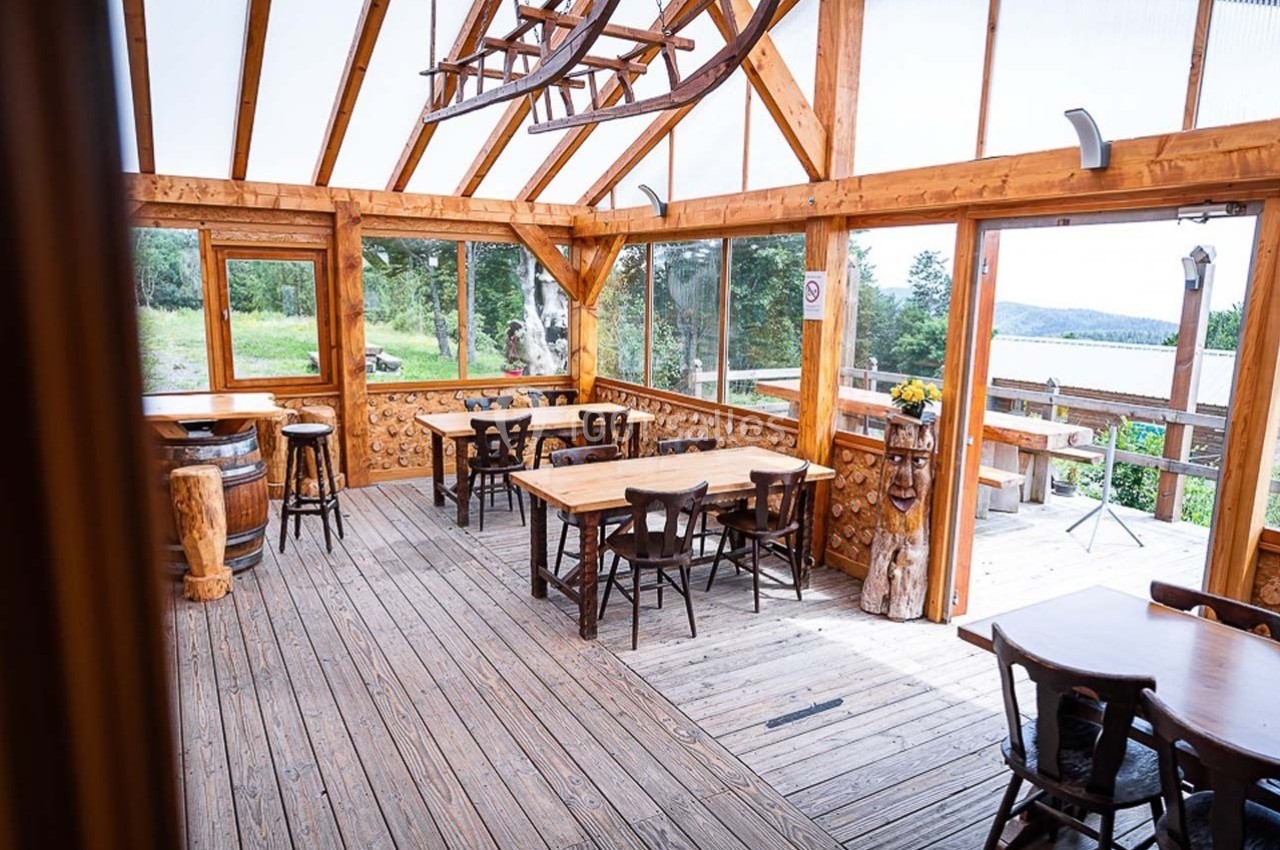 Salle en bois lumineuse avec de grandes fenêtres, des tables et des chaises, offrant une vue sur la nature environnante.