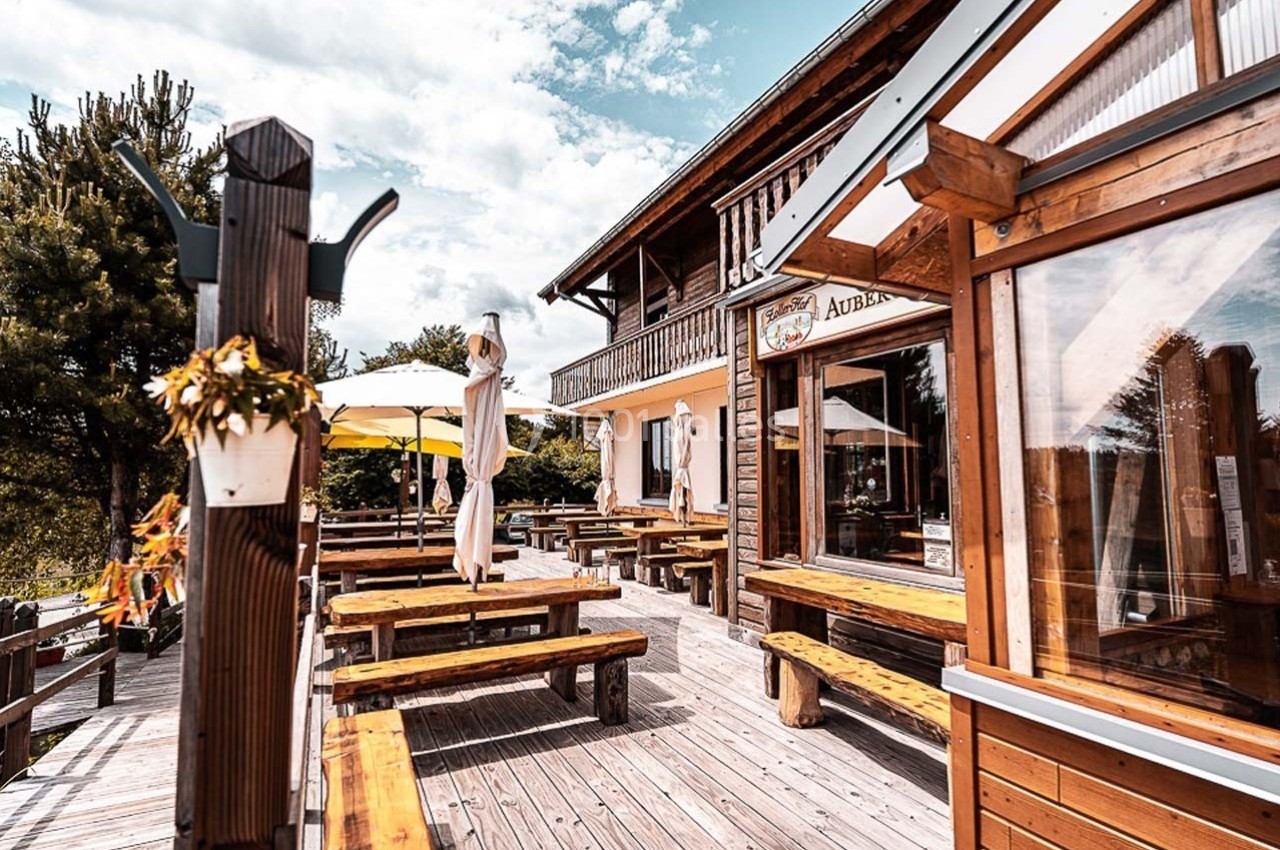 Terrasse en bois d'une auberge avec tables et parasols, entourée de végétation sous un ciel partiellement nuageux.