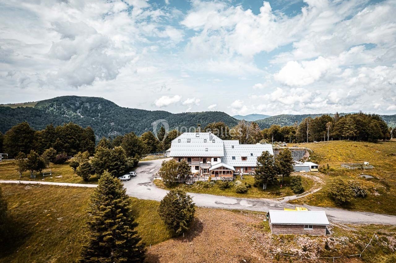 Vue aérienne d'un bâtiment entouré de prairies, d'arbres et de montagnes sous un ciel partiellement nuageux.