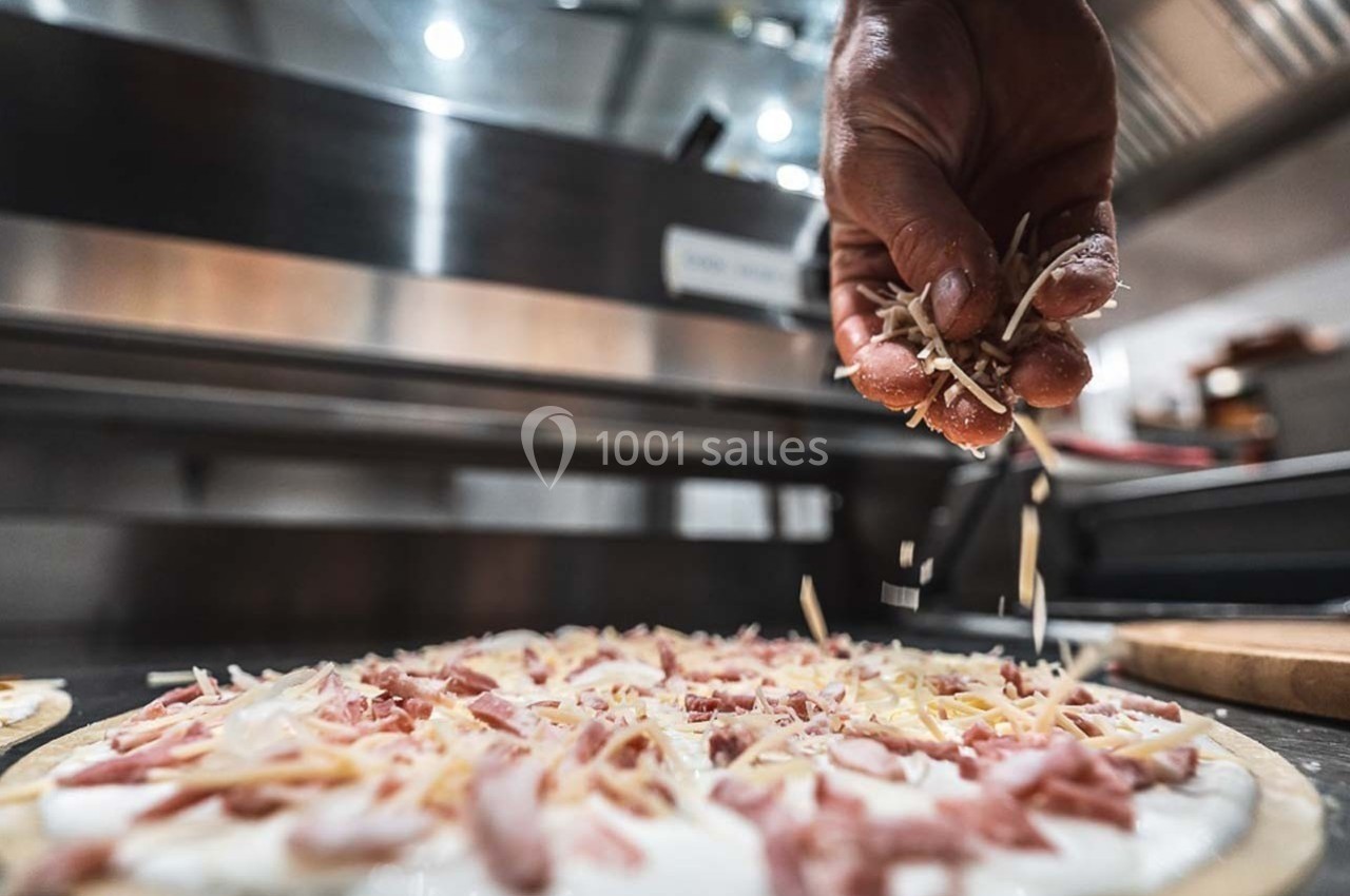 Une main saupoudre du fromage râpé sur une pizza garnie de jambon dans une cuisine professionnelle.