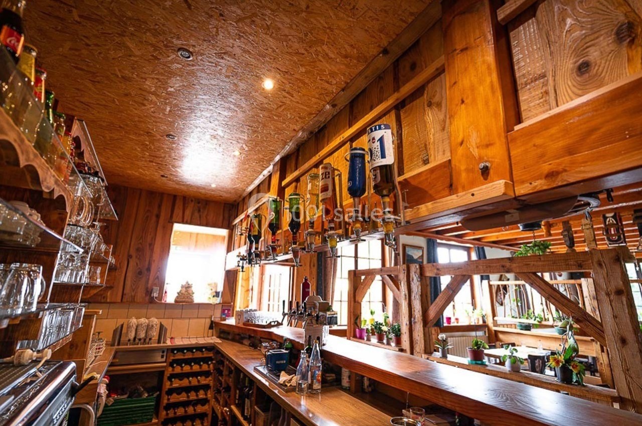 Intérieur d'un bar en bois avec des bouteilles suspendues, un comptoir et des étagères garnies de verres et de boissons.