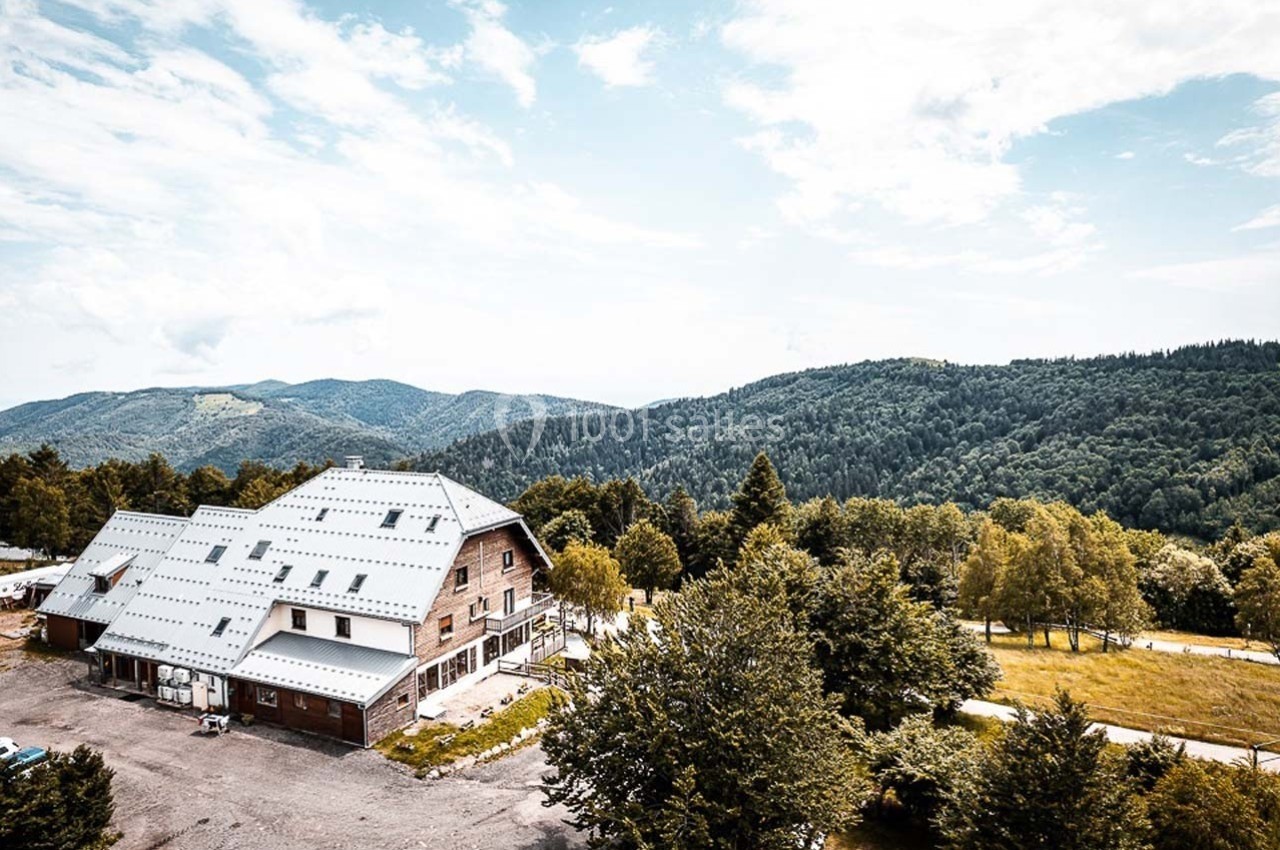 Bâtiment en bois avec toit métallique entouré de collines boisées sous un ciel partiellement nuageux.