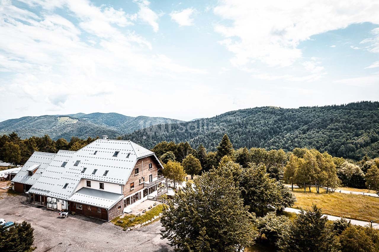 Bâtiment en bois avec toit métallique entouré de collines boisées sous un ciel partiellement nuageux.