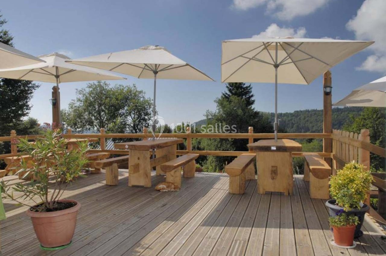 Terrasse en bois avec tables et bancs rustiques, entourée de plantes en pot et protégée par des parasols blancs.