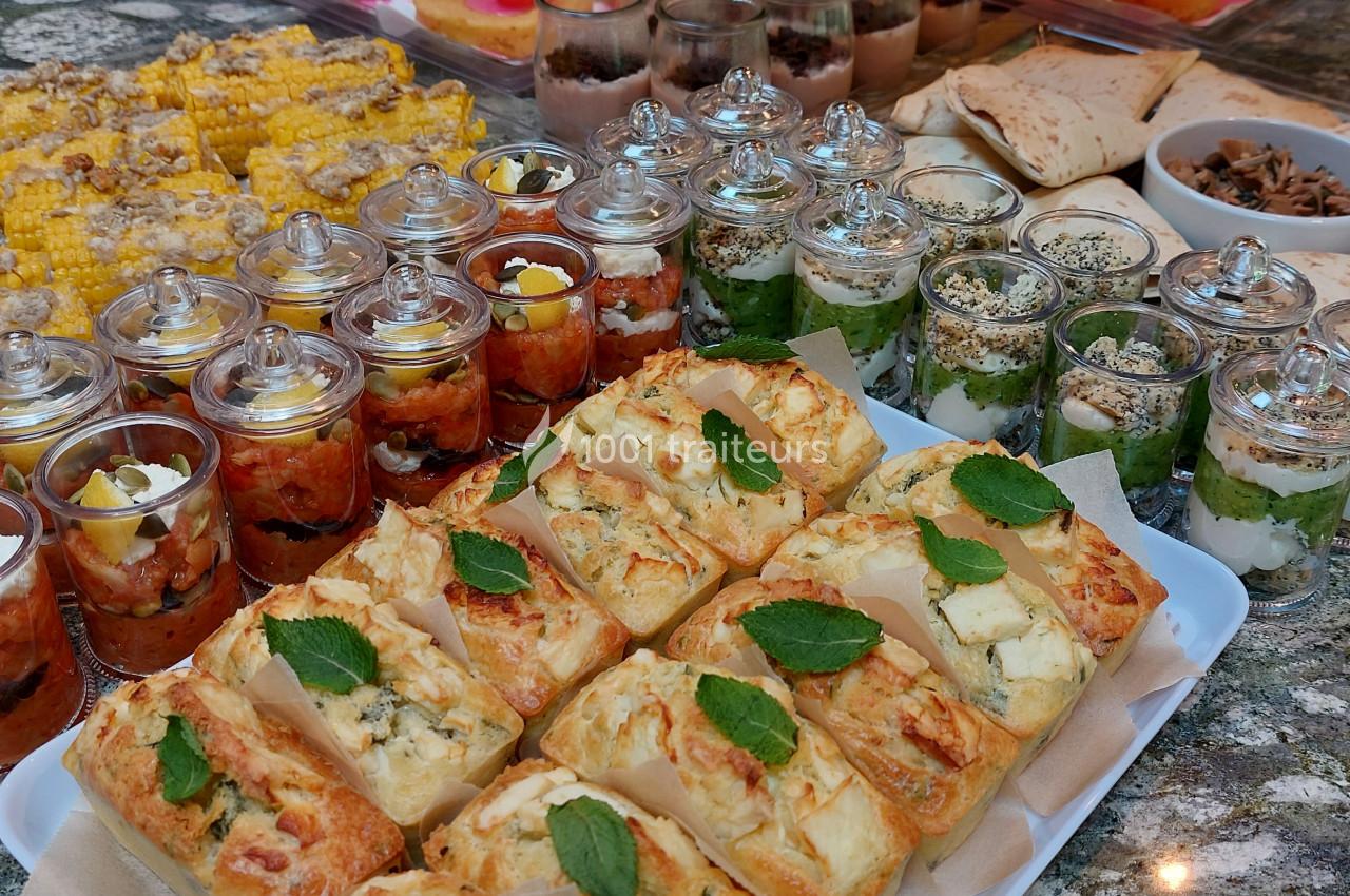 Plateau de buffet avec verrines salées, épis de maïs grillés, pains garnis et accompagnements variés sur une table en pierre.