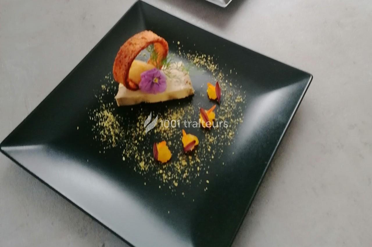 Dessert élégant sur assiette noire, décoré de fleurs comestibles, d'un biscuit courbé et de poudre colorée.