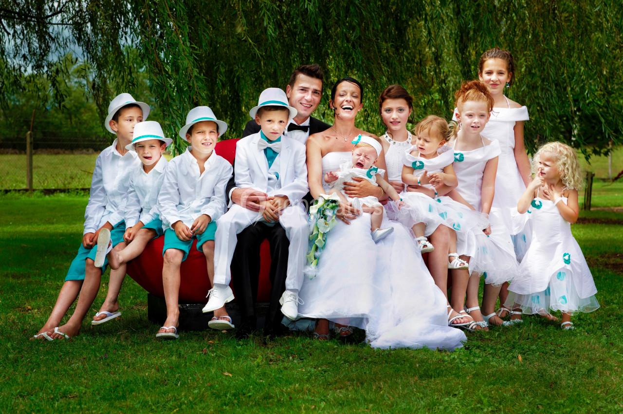 Un couple en tenue de mariage entouré d'enfants habillés en blanc et turquoise, posant dans un parc verdoyant.