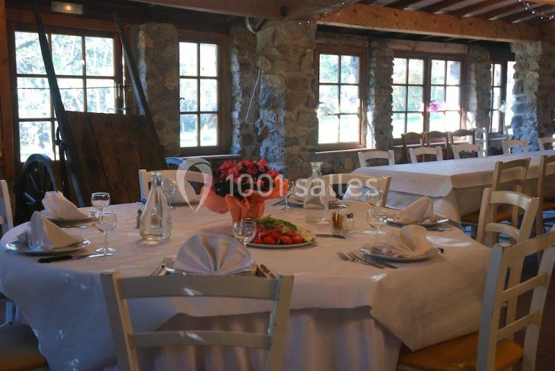 Table dressée avec nappes blanches, assiettes, verres et un plat de crudités, dans une salle rustique avec fenêtres en bois.