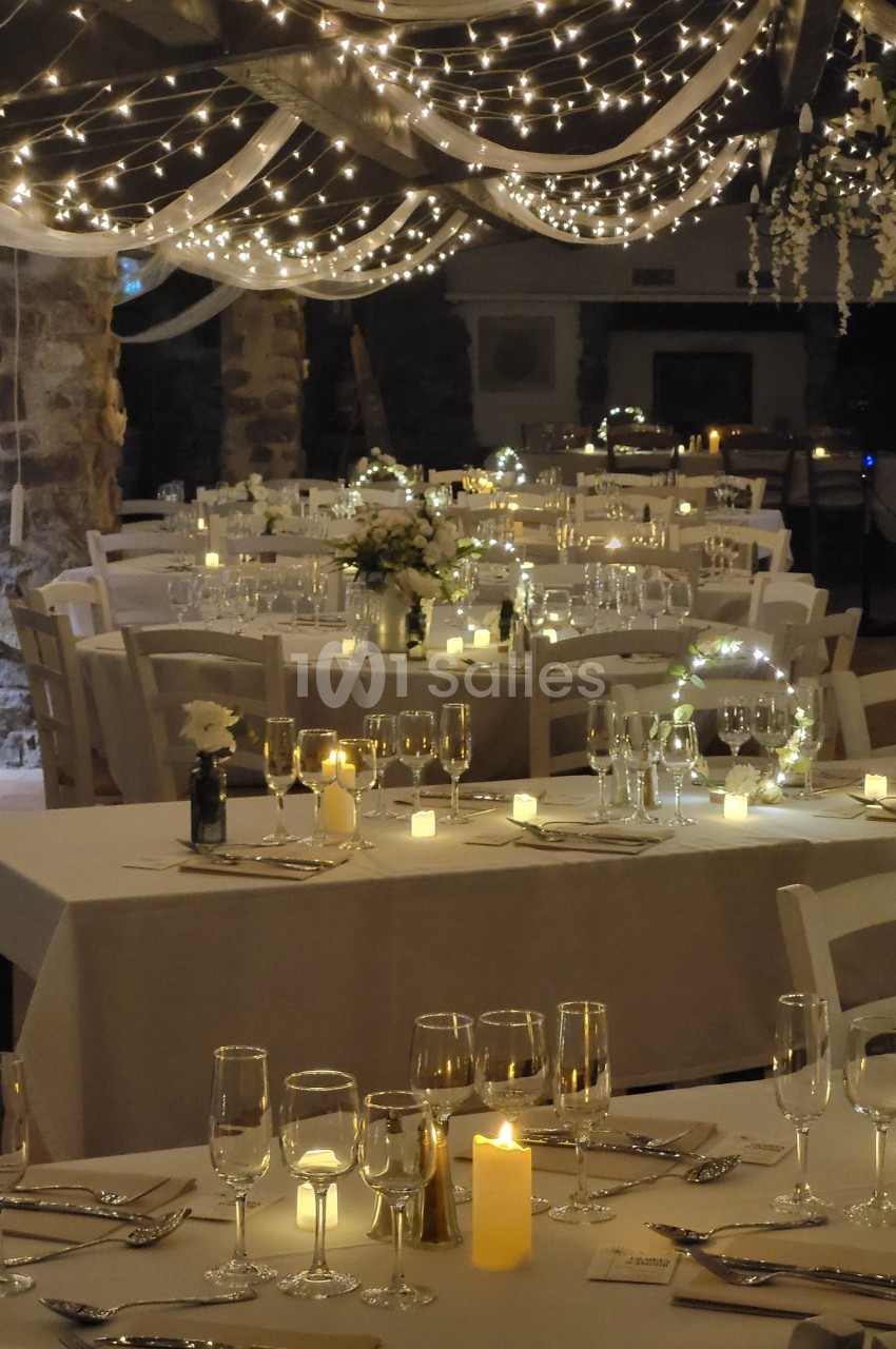 Salle de réception décorée avec des guirlandes lumineuses, des tables dressées avec nappes blanches et des bougies allumées.
