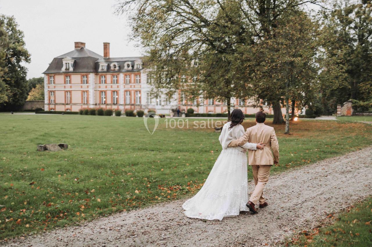 Un couple en tenue de mariage marche sur un chemin gravillonné devant un château entouré de verdure.