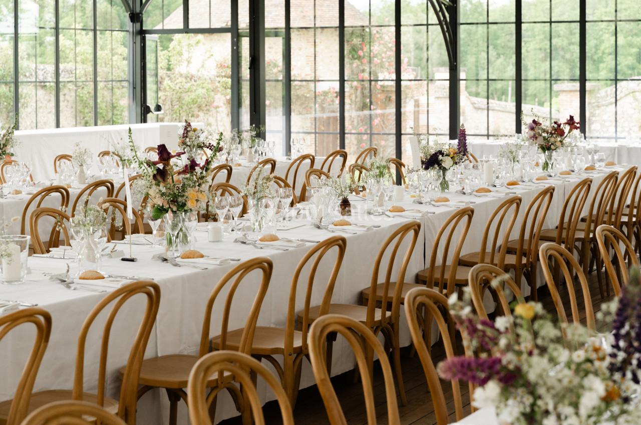 Salle de réception lumineuse avec de longues tables dressées, décorées de fleurs et entourées de chaises en bois.