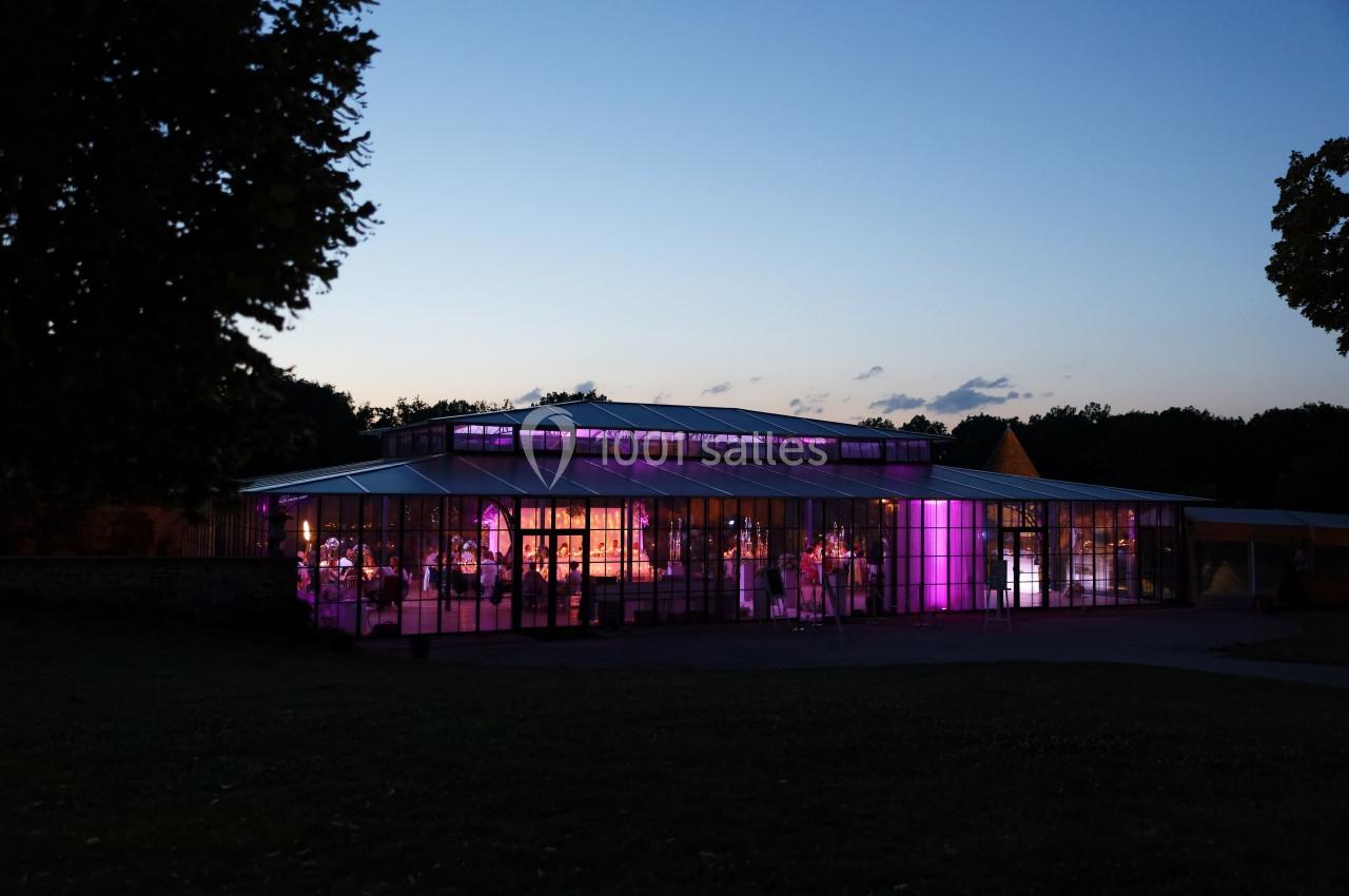 Bâtiment en verre illuminé de lumières violettes à la tombée de la nuit, entouré de végétation.