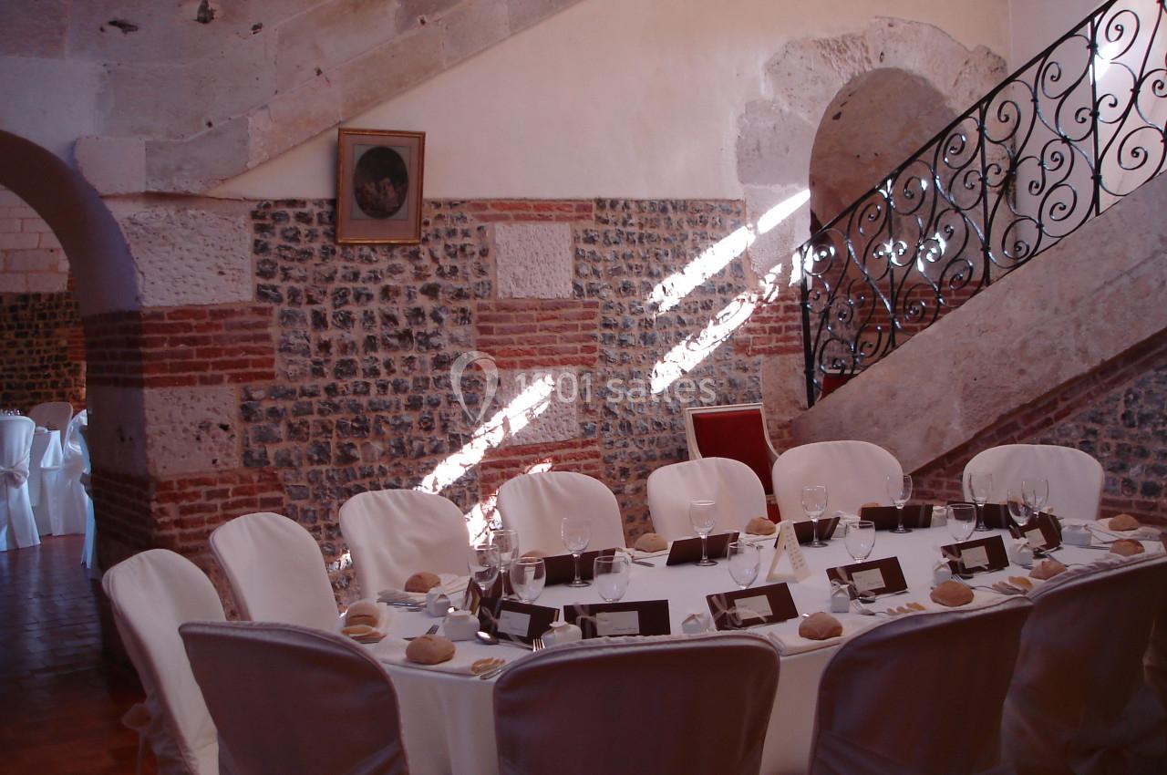 Table élégamment dressée avec nappes blanches, couverts et porte-noms, dans une salle en pierre avec escalier en fer forgé.
