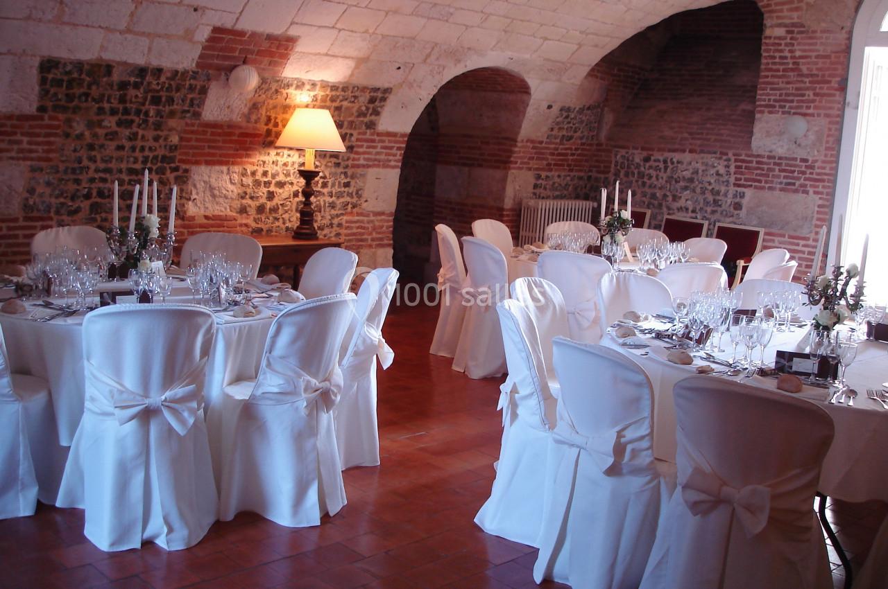 Salle voûtée en pierre aménagée pour un banquet, avec tables dressées et chaises recouvertes de housses blanches.