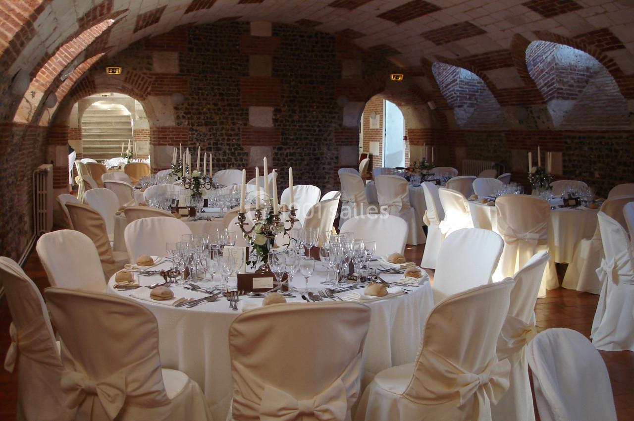 Salle voûtée en briques aménagée pour un événement avec des tables rondes dressées et des chaises couvertes de housses…