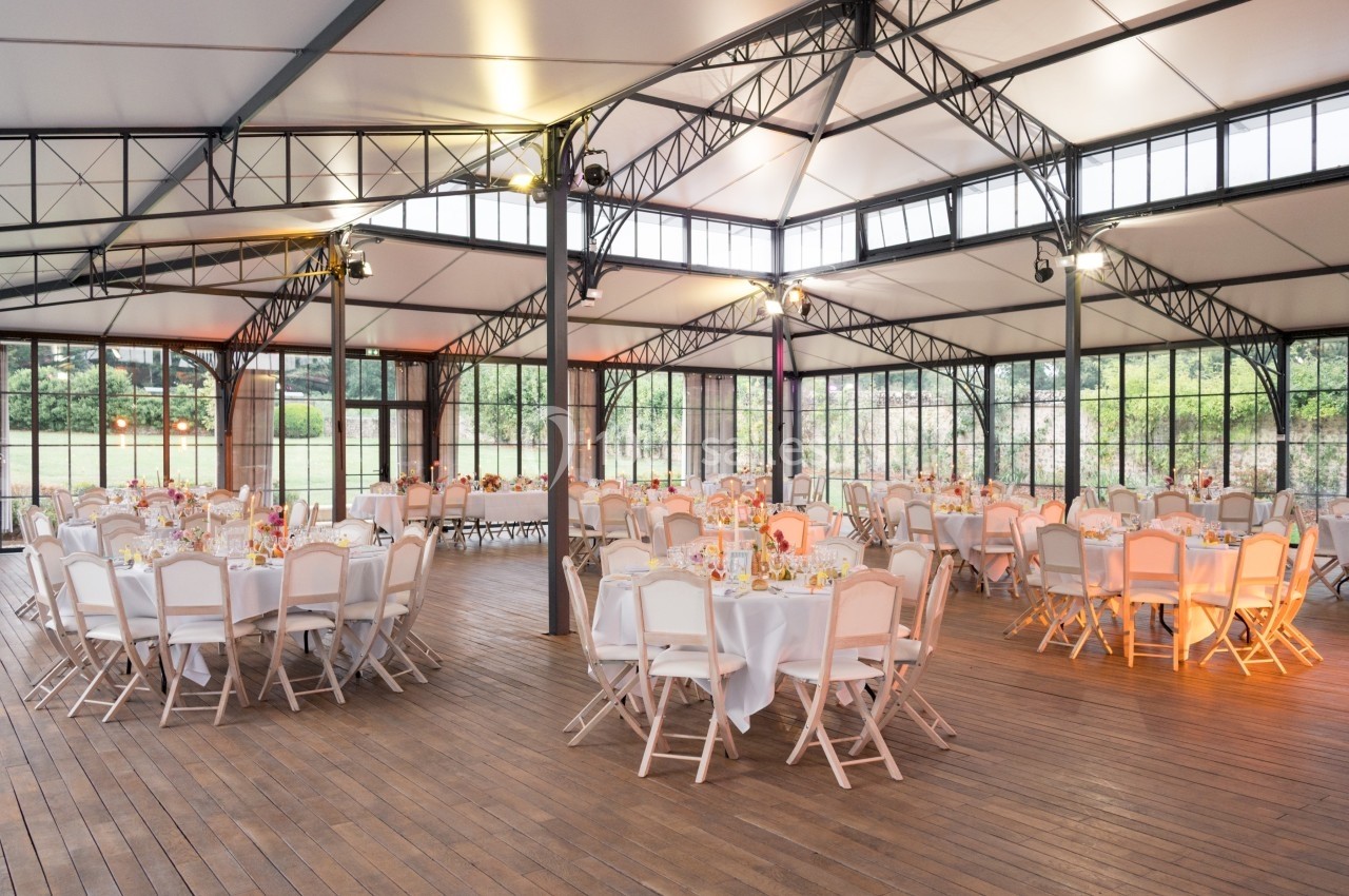 Salle de réception lumineuse avec tables rondes dressées et chaises blanches, sous une structure métallique vitrée.