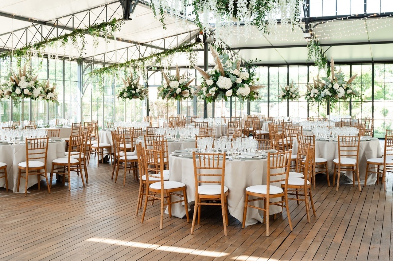 Salle de réception lumineuse avec tables rondes dressées, chaises en bois et décorations florales suspendues.