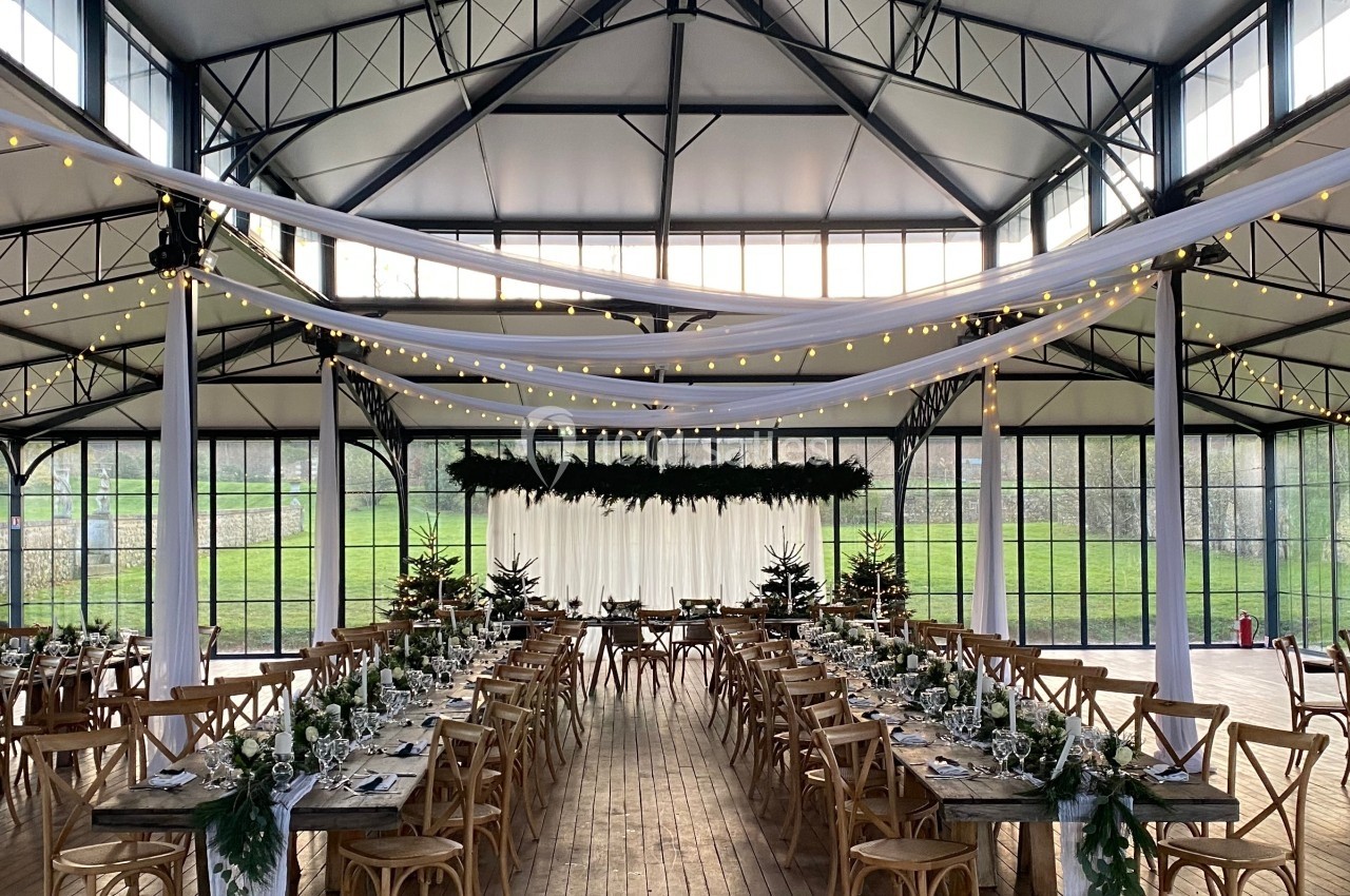 Salle de réception lumineuse avec tables en bois décorées de verdure et guirlandes lumineuses suspendues.