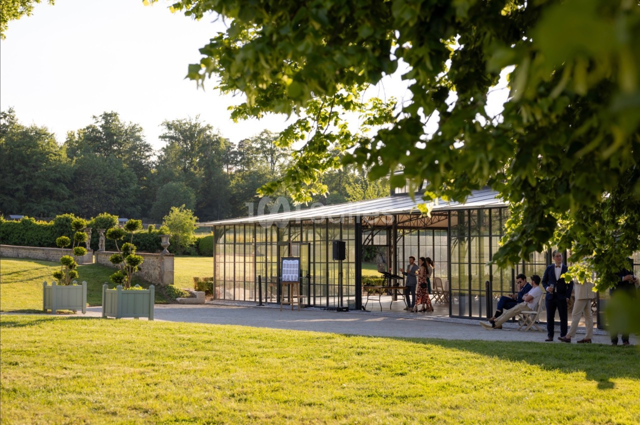 Serre vitrée entourée de verdure, avec des personnes à l'intérieur et à l'extérieur dans un cadre champêtre.