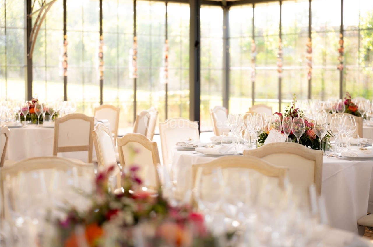 Salle lumineuse avec tables décorées de nappes blanches, bouquets de fleurs et verres disposés pour un événement.