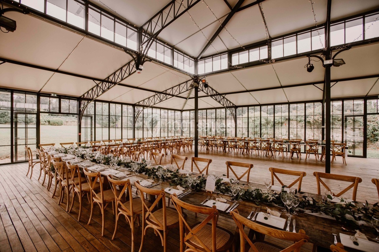 Grande salle lumineuse avec charpente métallique, tables en bois alignées et décorées de verdure, vue sur l'extérieur…