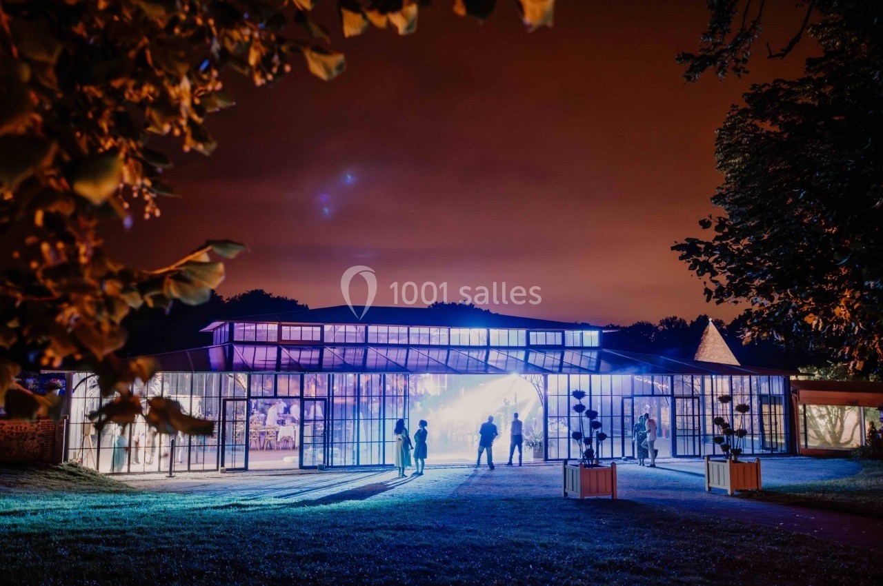 Serre illuminée de nuit avec des silhouettes de personnes à l'extérieur, entourée d'arbres et d'un ciel orangé.