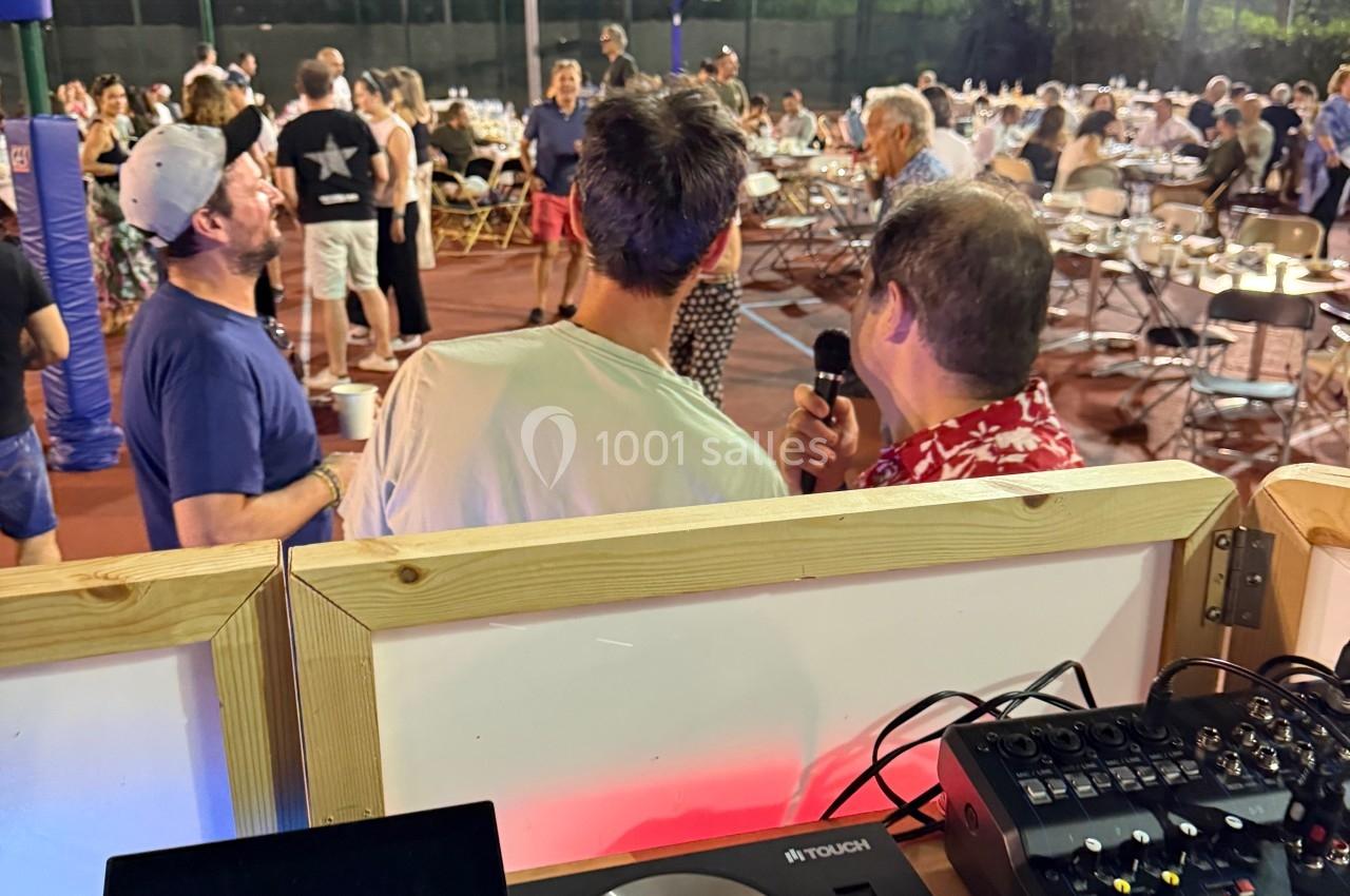 Des personnes discutent sur un terrain de sport en plein air, avec des tables installées et une console de DJ au premier…