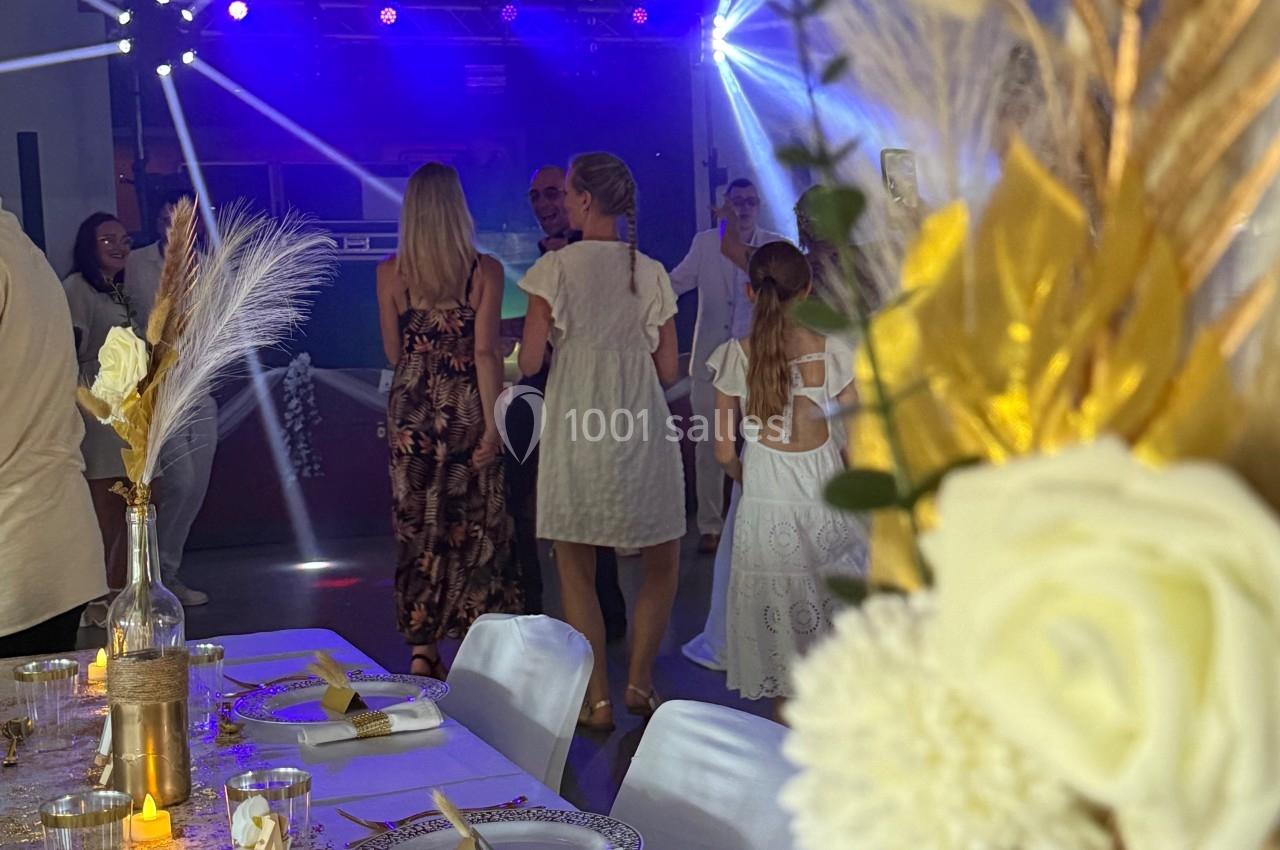 Salle décorée pour un événement avec table élégante au premier plan et invités dansant sous des jeux de lumière en arrière…