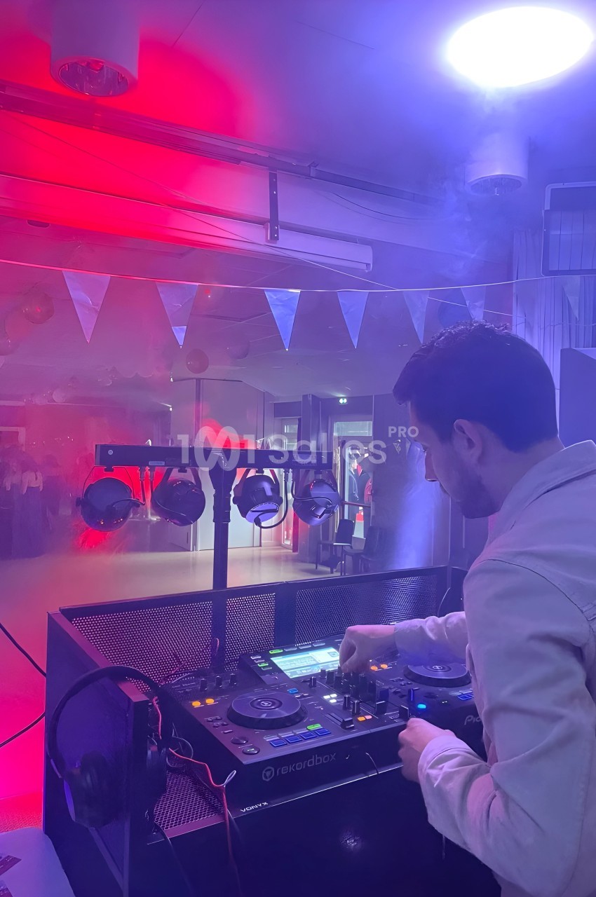 Un DJ mixe sur une console dans une salle éclairée par des lumières rouges et violettes, avec des invités en arrière-plan.