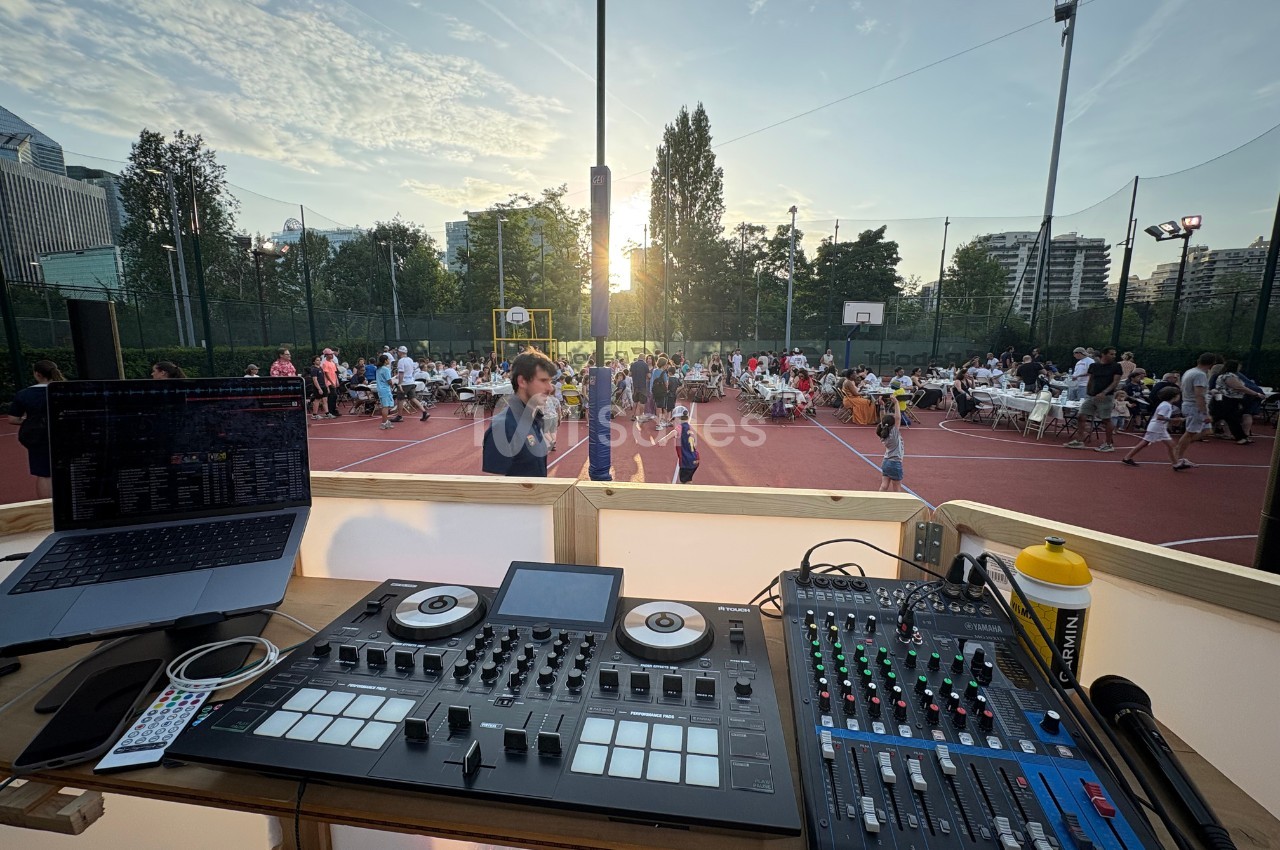 Vue d'une table de mixage et d'un ordinateur surplombant un terrain de sport avec des tables et des convives en plein air.