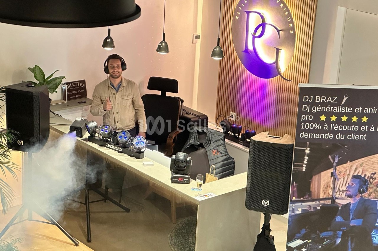 Un DJ derrière une table de mixage avec éclairage et matériel audio, dans un espace moderne avec un panneau lumineux.