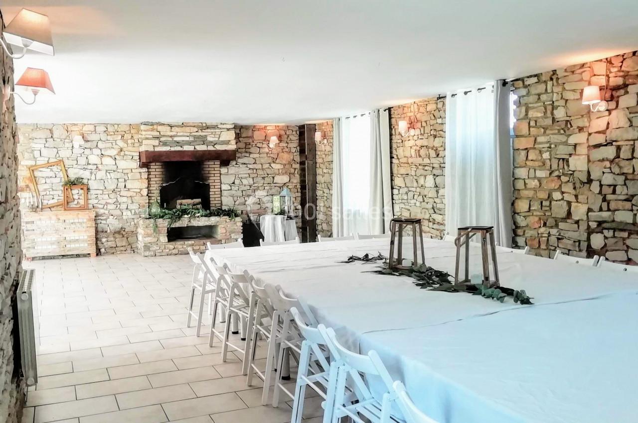 Salle en pierre avec des tables blanches alignées, des chaises pliantes et une cheminée en arrière-plan.
