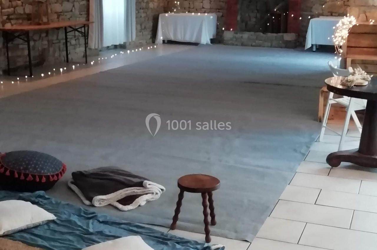 Salle lumineuse avec murs en pierre, tapis, coussins au sol, tables en fond et cheminée décorative.