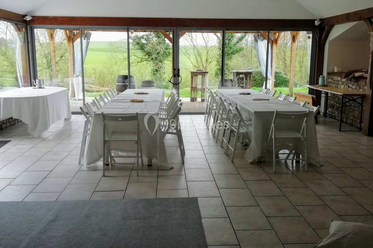 Salle lumineuse avec grandes baies vitrées, tables et chaises blanches disposées pour un repas, vue sur un jardin verdoyant.