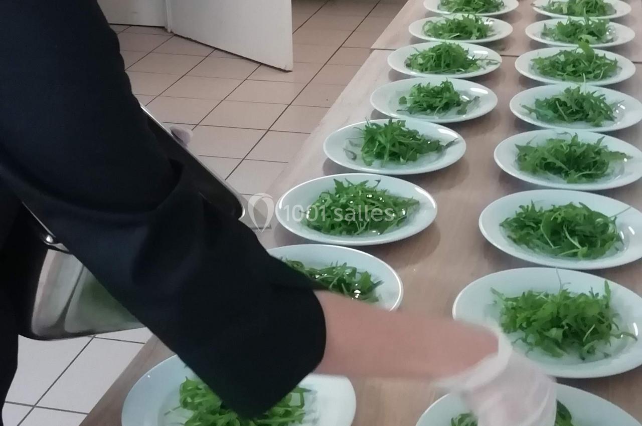 Une personne dispose des assiettes garnies de salade verte sur une longue table dans une cuisine.