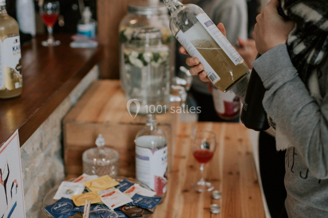 Une femme verse une boisson dans un verre sur un comptoir en bois, entourée de verres et de sachets de thé.