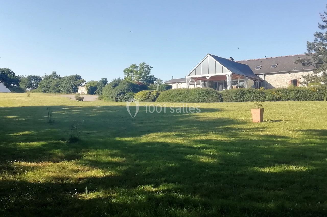 Grande pelouse verdoyante avec une maison en pierre et un toit en ardoise entourée de haies et d'arbres.