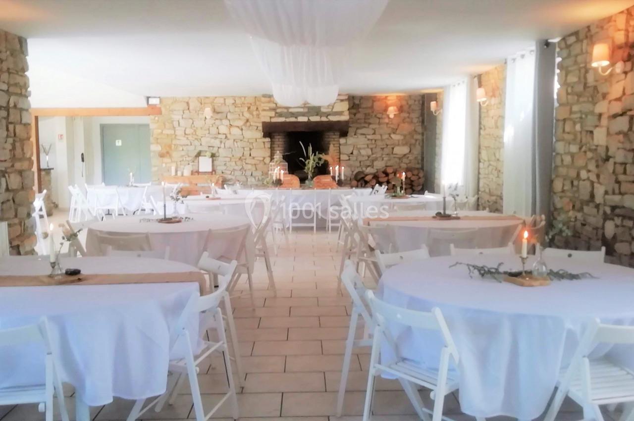Salle de réception lumineuse avec murs en pierre, tables rondes dressées de nappes blanches et décorations simples.