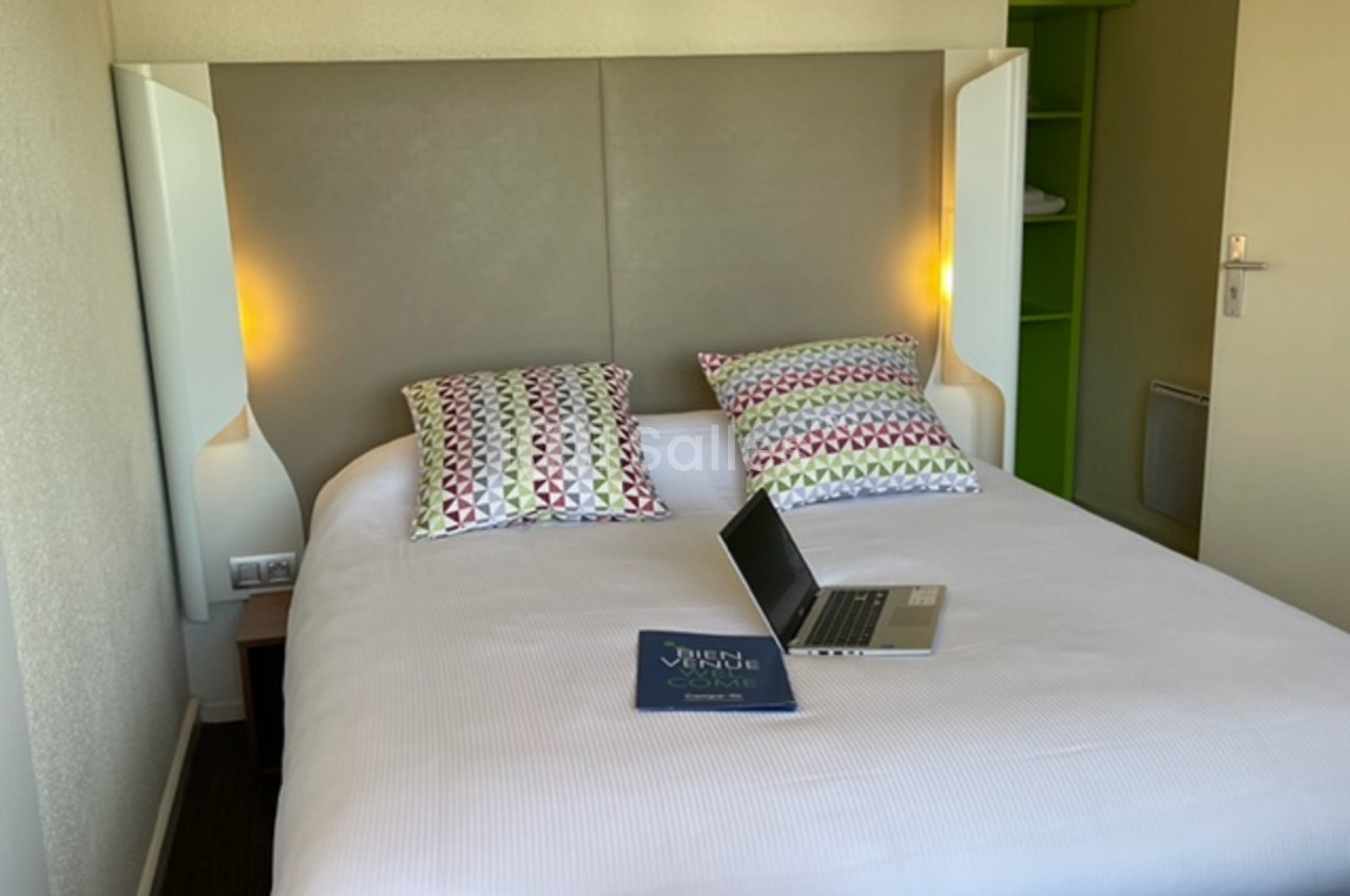 Lit double avec draps blancs, deux coussins à motifs colorés, un ordinateur portable et un livret posés dessus.