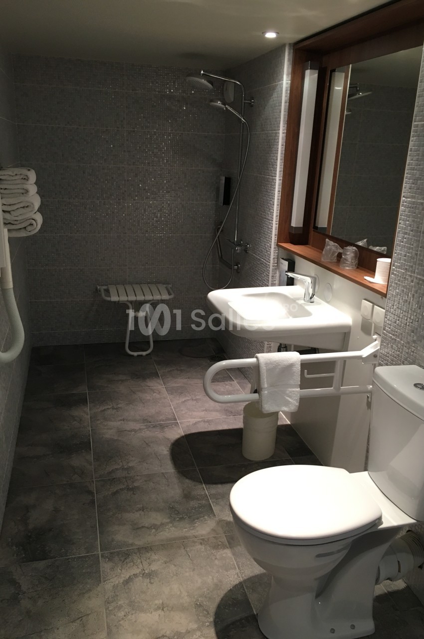 Salle de bain accessible avec douche à l'italienne, barre d'appui, lavabo, toilettes et carrelage gris.