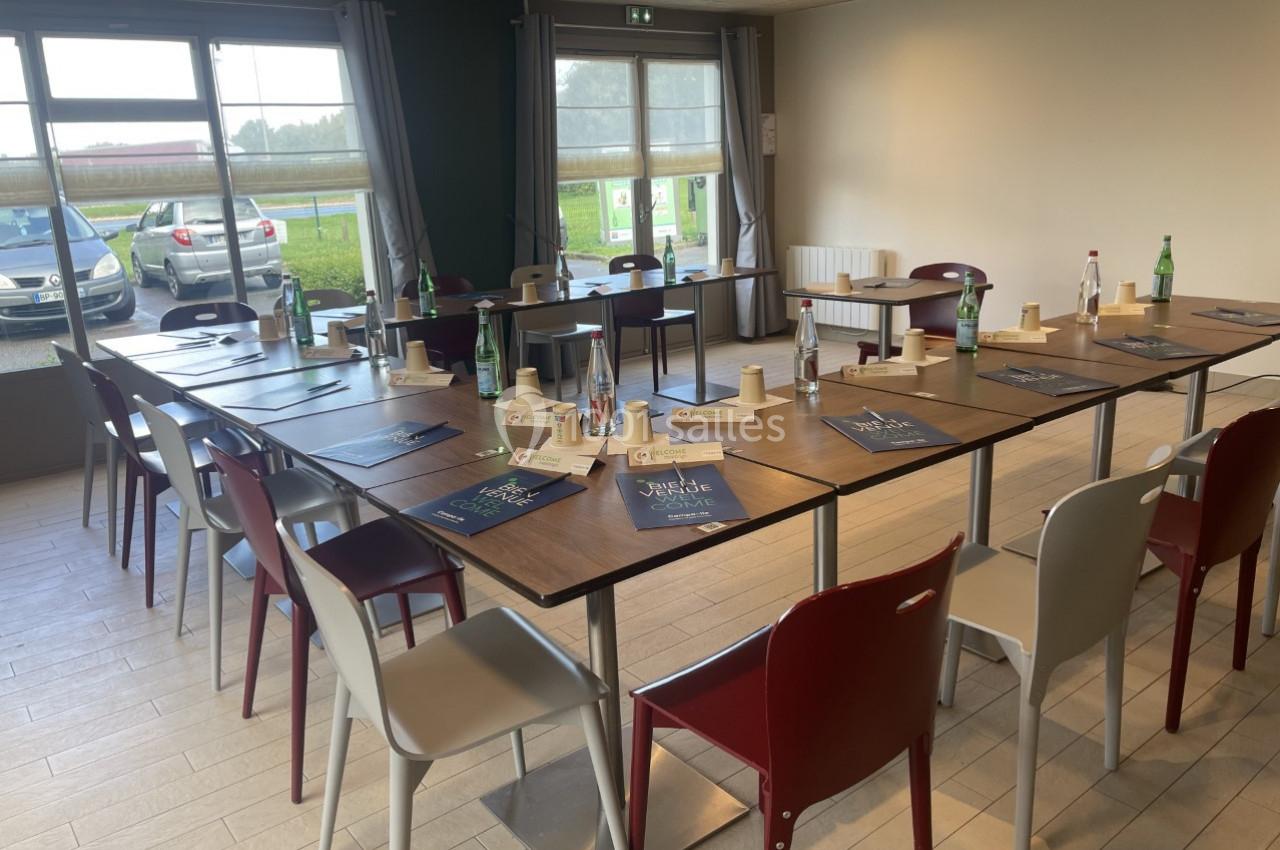 Salle de réunion lumineuse avec tables disposées en U, chaises colorées, documents et bouteilles d'eau sur les tables.