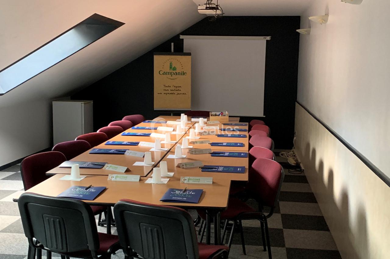 Salle de réunion sous pente avec table rectangulaire, chaises rouges, projecteur, écran blanc et lumière naturelle.