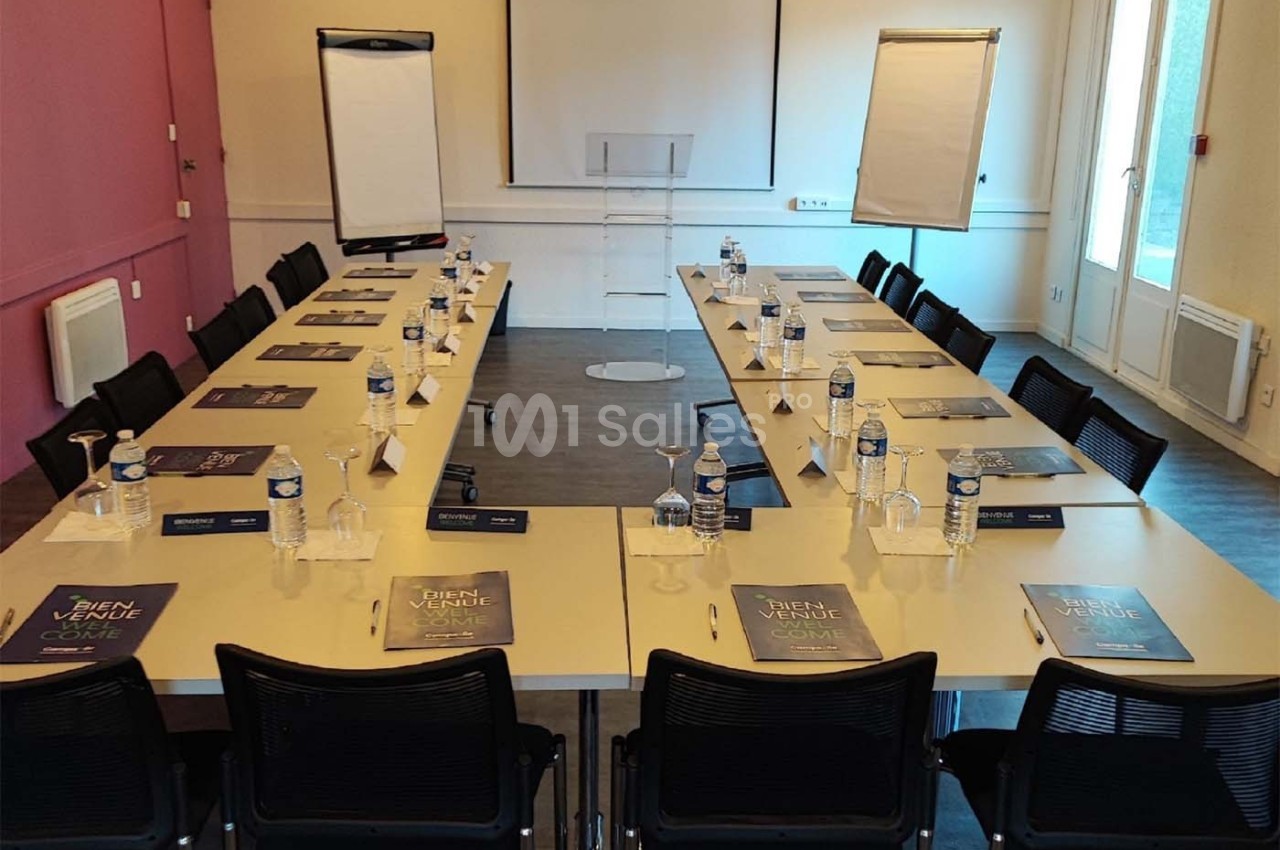 Salle de réunion équipée de tables en U avec chaises, blocs-notes, bouteilles d'eau, paperboards et écran de projection.