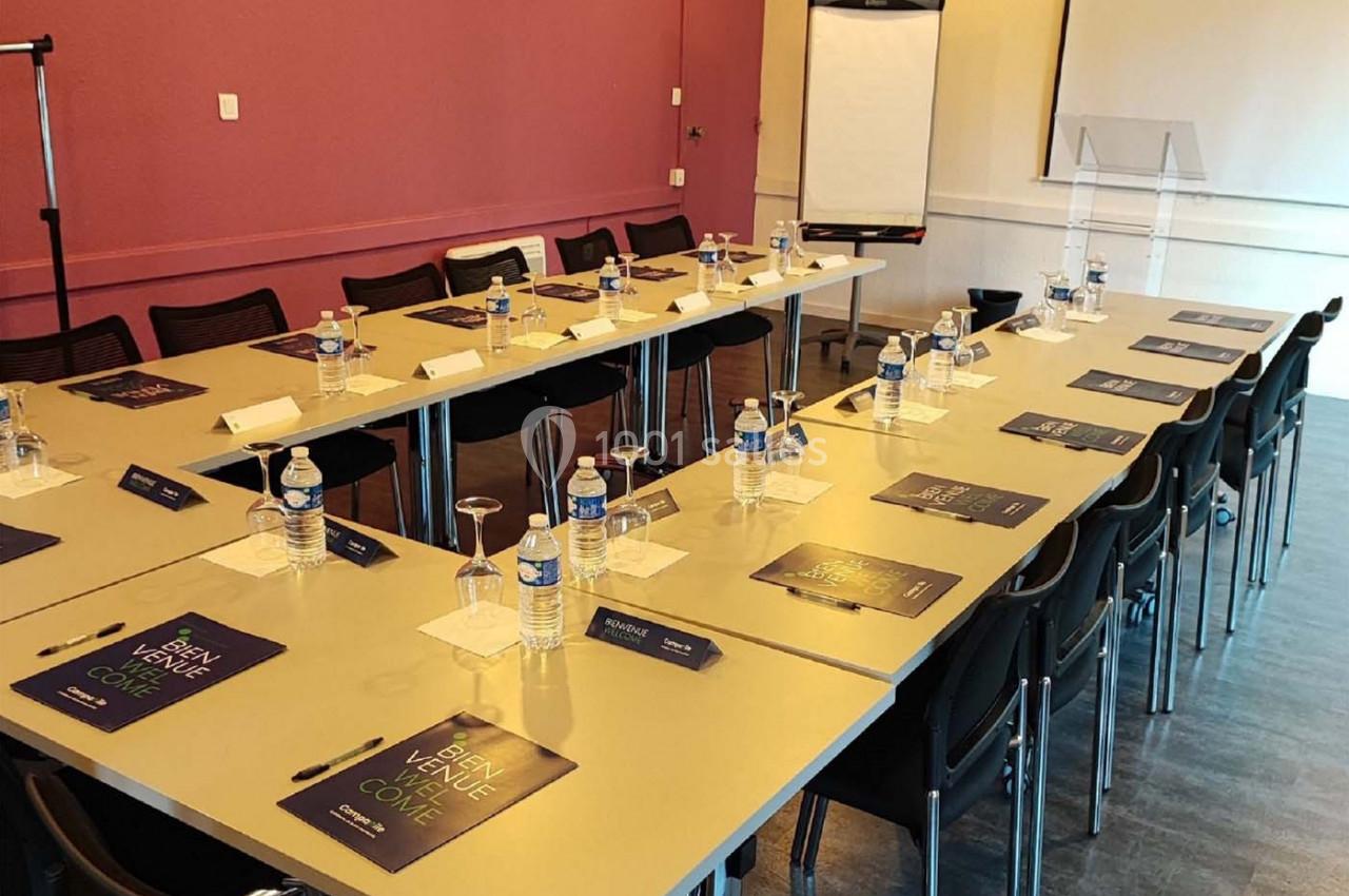 Salle de réunion avec tables en U, chaises, bouteilles d'eau, documents, un paperboard et un pupitre transparent.