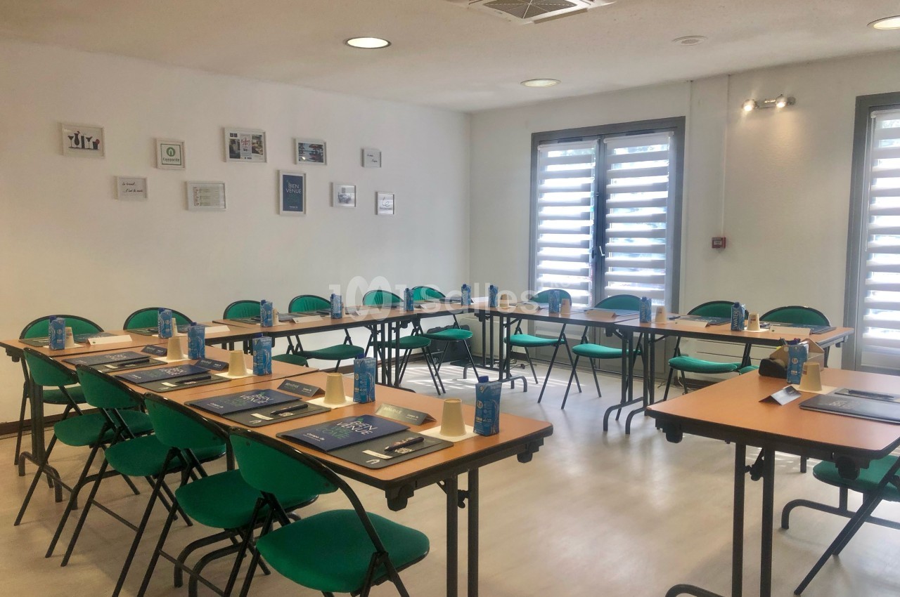 Salle de réunion lumineuse avec tables disposées en U, chaises vertes et matériel de travail sur chaque place.