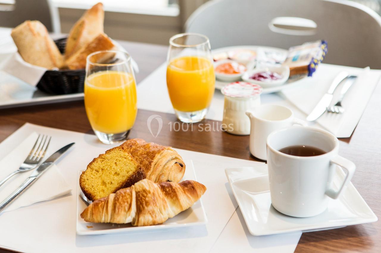 Petit-déjeuner avec croissants, tranche de gâteau, jus d'orange, café et condiments sur une table dressée.